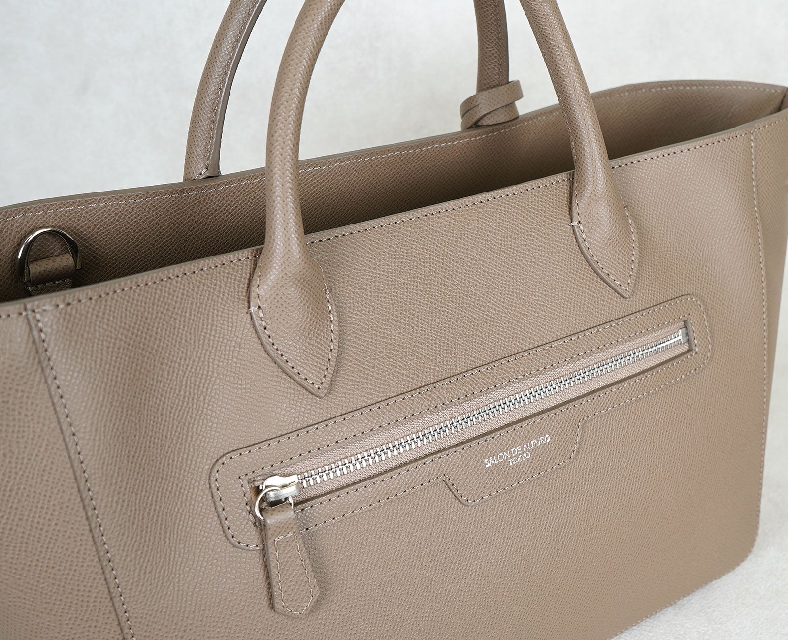 ファスナーまわりまで洗練された裏面デザイン。
木の葉型にカットされたハンドル根元や、〈サロンドアルファード〉のロゴを箔押ししたディテールが、上質なアクセントに。